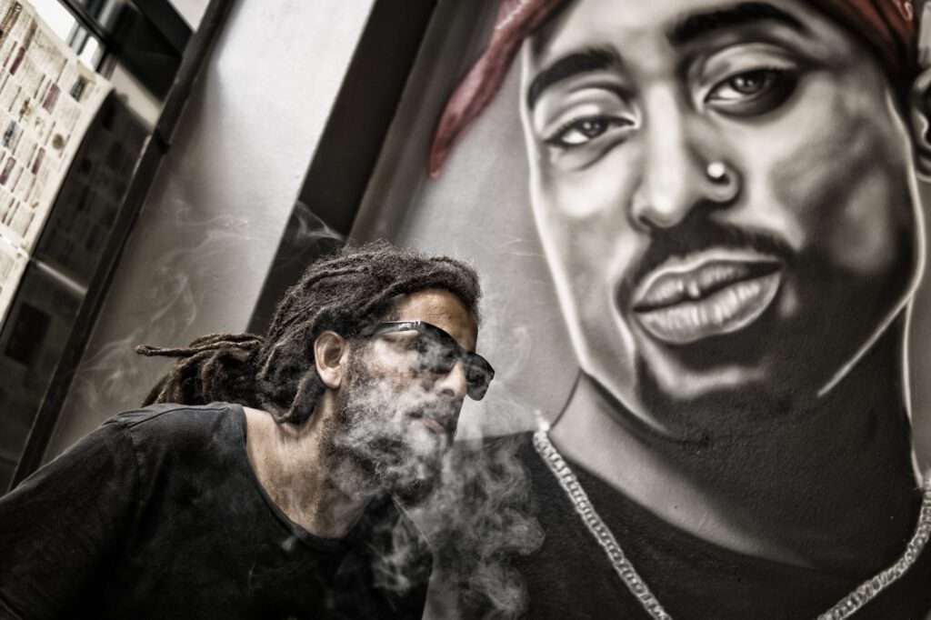 man with dreadlocks and sunglasses poses near tupac shakur portrait